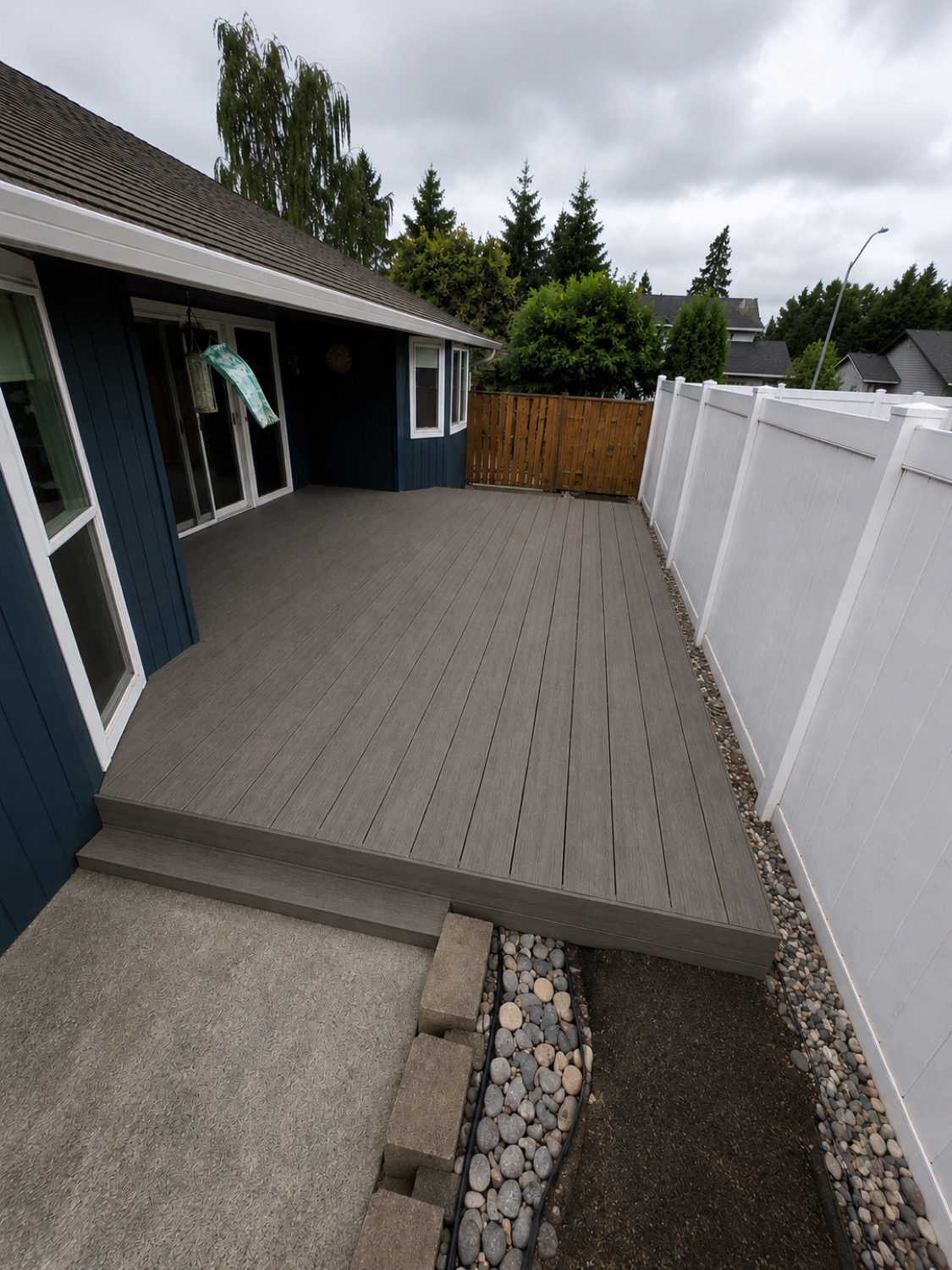 Ground-level deck beside a blue house and white privacy fence